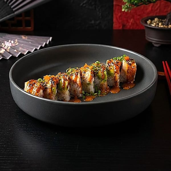 Sushi rolls with sauce and toppings on a dark plate, with chopsticks and a fan.