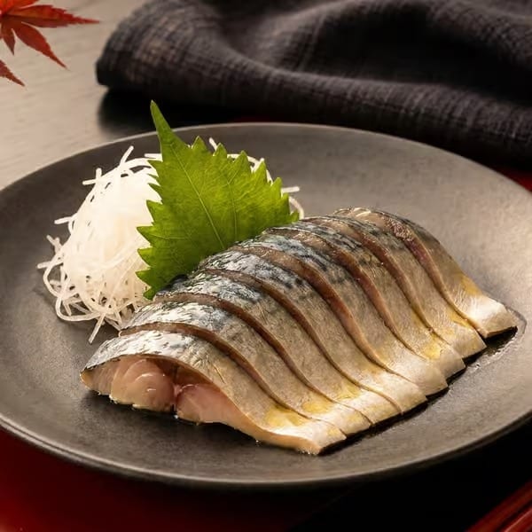 Sliced mackerel sashimi with shredded daikon and a green shiso leaf on a dark plate.