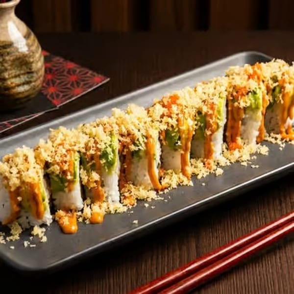 Sushi roll with avocado, orange sauce, and crispy toppings on a dark rectangular plate.